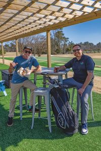 Two men are sitting on barstools at a high table outdoors under a wooden pergola. One is holding food, and the other a drink. A golf bag is beside them. The scene is bright and sunny, with a golf course and trees in the background.
