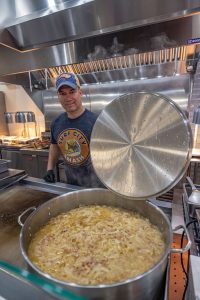 A man in a baseball cap and Surf City Mash t-shirt is standing in a commercial kitchen. He is holding the lid of a large metal pot filled with a simmering mixture of onions and sauce. The kitchen has industrial equipment and a vent hood.