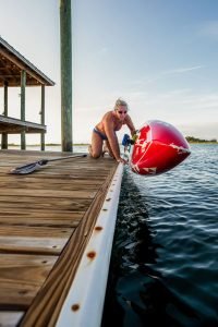 A woman in swimwear and pink goggles lifts a red kayak from the water onto a wooden dock beside a lakeside structure, with a paddle resting on the dock.