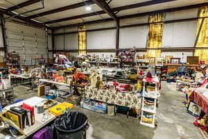 A large indoor garage sale with tables covered in various secondhand items, including toys, decorations, electronics, and household goods, set up in a spacious warehouse with yellow curtains on the windows.