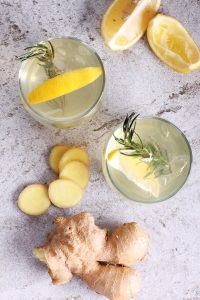 Two glasses of lemon-ginger drink with rosemary sprigs, surrounded by fresh ginger root, ginger slices, and lemon wedges on a light gray surface.