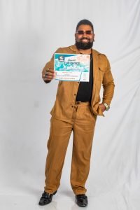 A man in a brown suit and sunglasses smiles while holding a "Top Choice Awards" certificate, standing against a plain white backdrop.