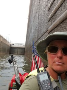 A person wearing sunglasses and a sunhat sits in a kayak with an American flag, paddling through a tall, narrow canal or lock with high concrete walls and water below.