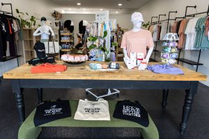 Clothing boutique interior with folded shirts, hats, and accessories on a wooden table; tote bags on a bench and racks of clothes in the background; bright, organized, and welcoming retail space.